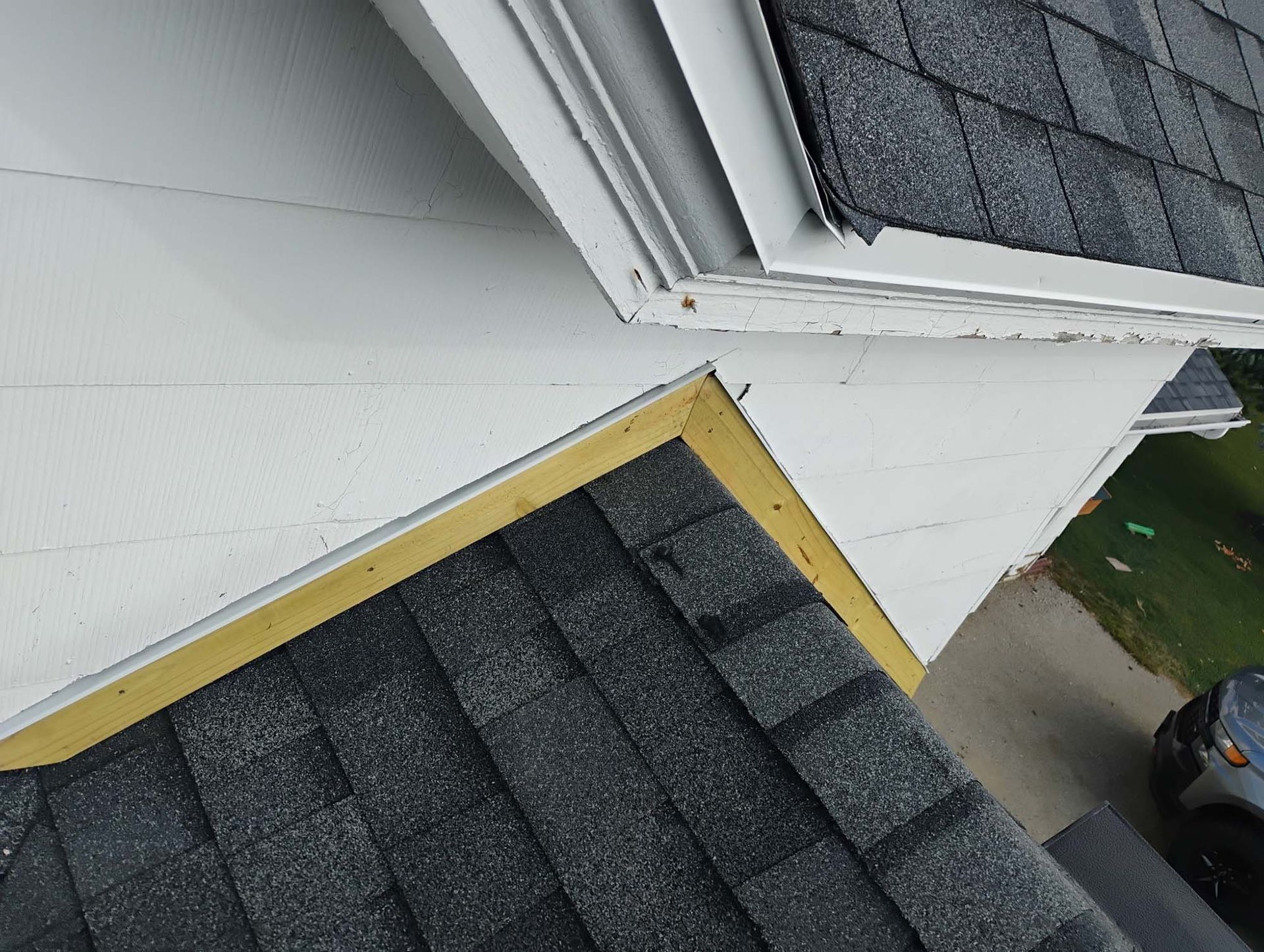 Overhead view of a roof intersection with dark shingles, white siding, and a wooden trim.