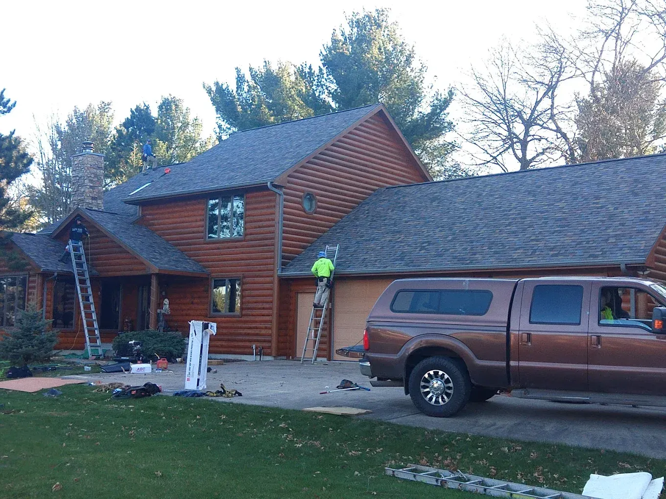 Workers on a brown house roof, a brown truck in the driveway. Trees in background.