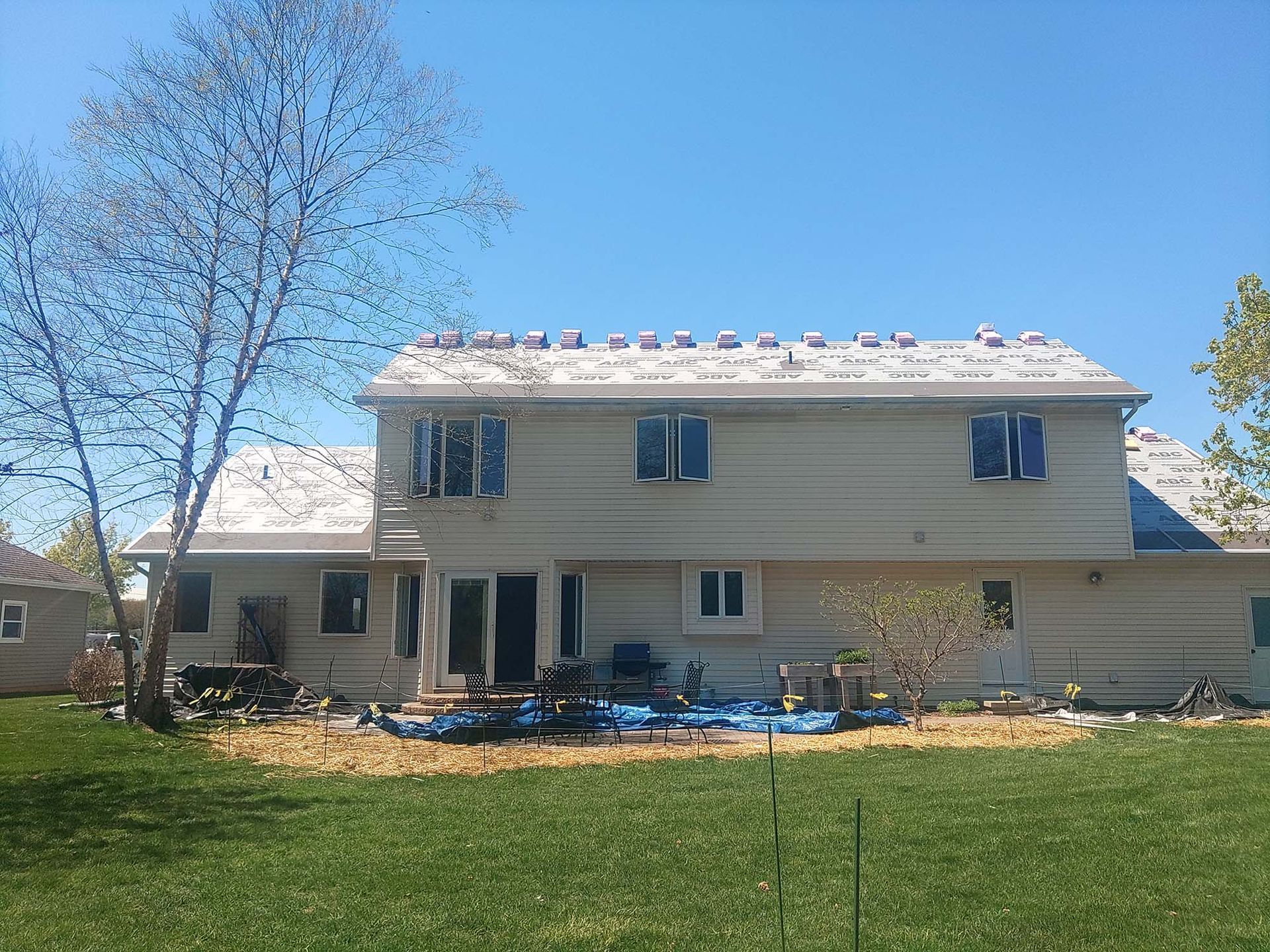 Back view of a two-story beige house with roof repair underway, sunny day.