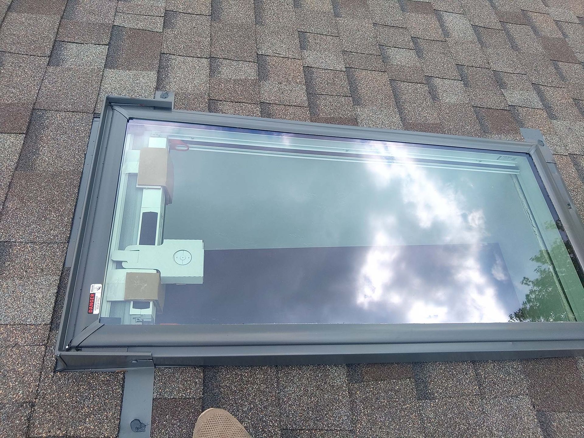 Skylight on a shingled roof, showing the glass and frame. Cloudy sky reflects in the glass.
