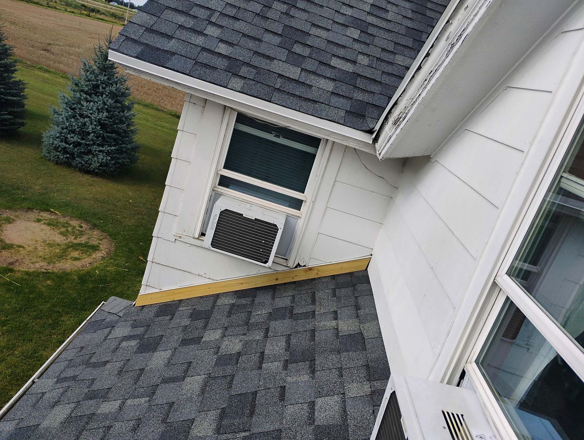 Overhead view of a house with gray shingles, white siding, a window-mounted air conditioner, and a green lawn.