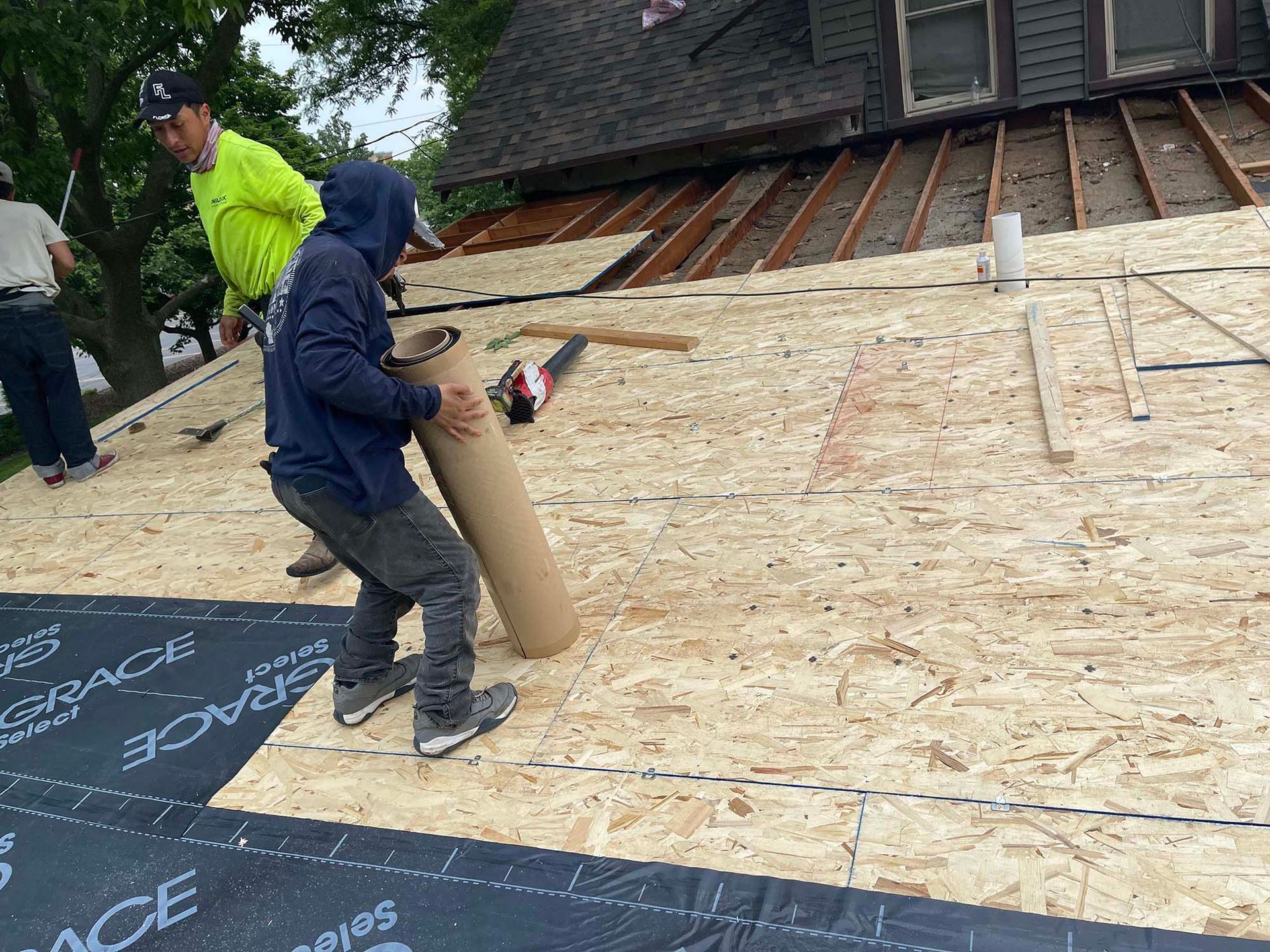 Roofers installing materials on a house roof. One holds a roll while others work on the wooden base.