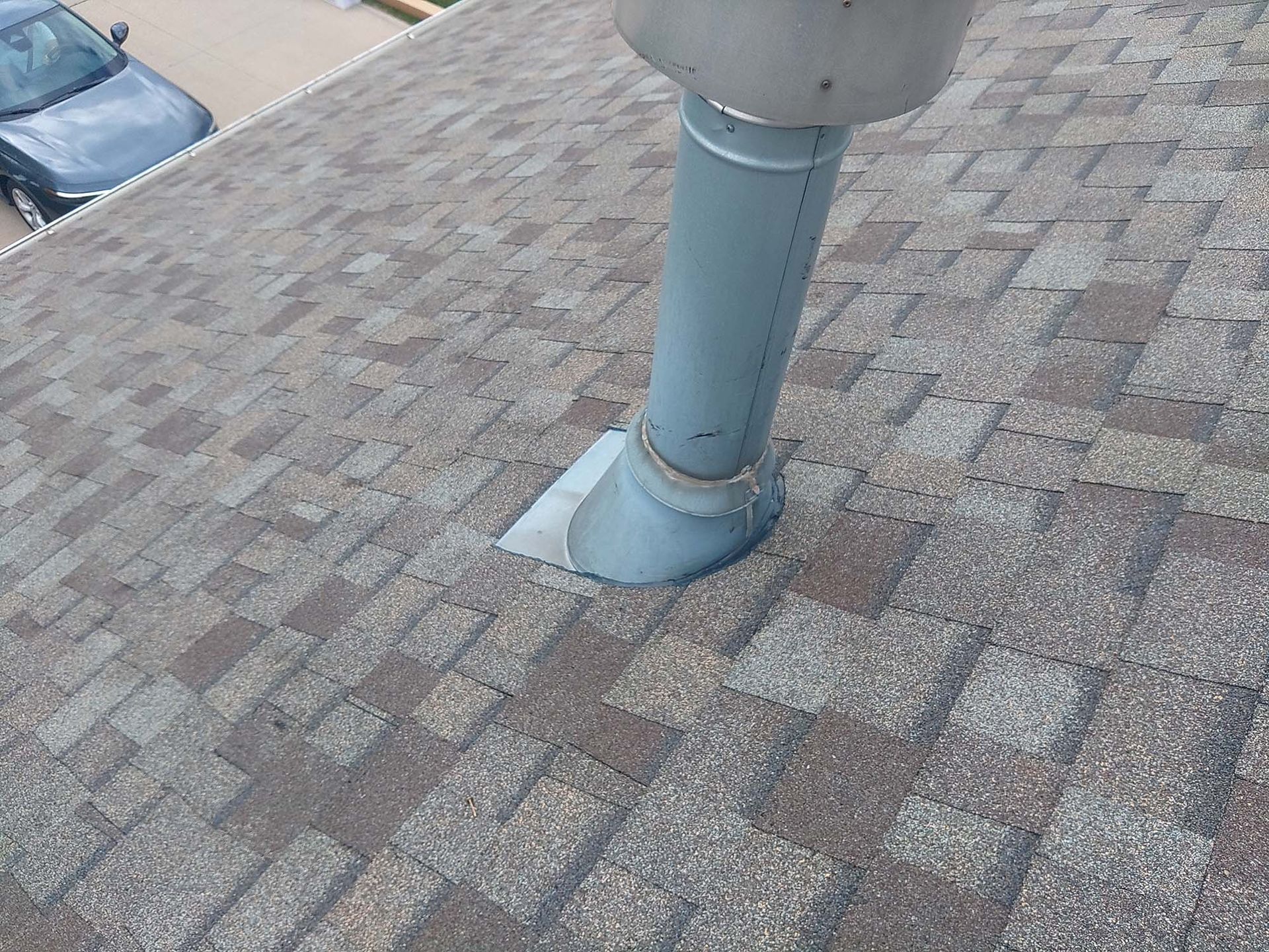 Gray vent pipe rising from a shingled roof. A car is visible on the left.