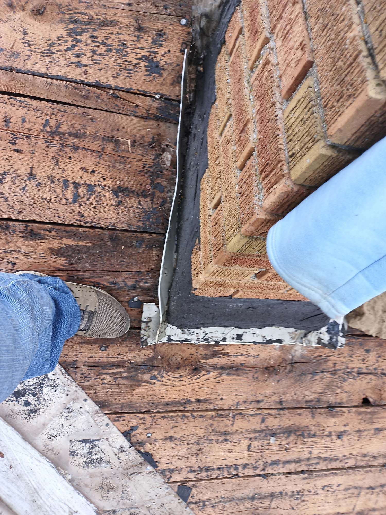 A person standing on a weathered wooden roof next to a brick wall with sealant.