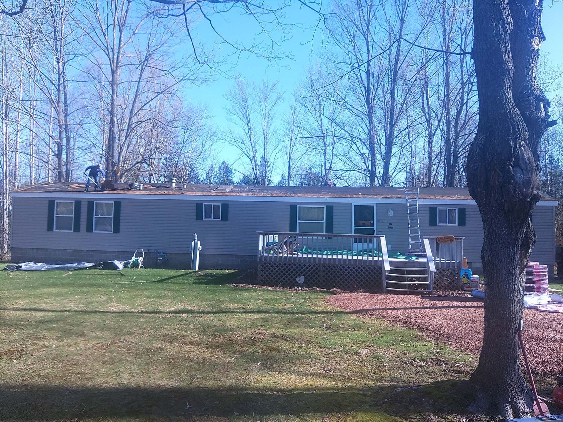 A gray mobile home with a deck and surrounding yard, trees in the background, blue sky.