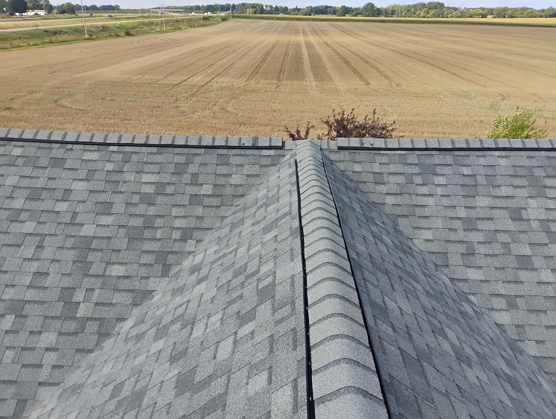 Gray shingled roof ridge with a brown field in the background under a blue sky.
