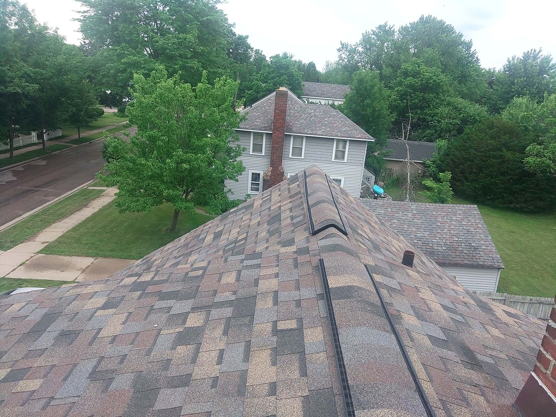 Overhead view of a house roof, foreground. A street, trees, and another house are in the background.