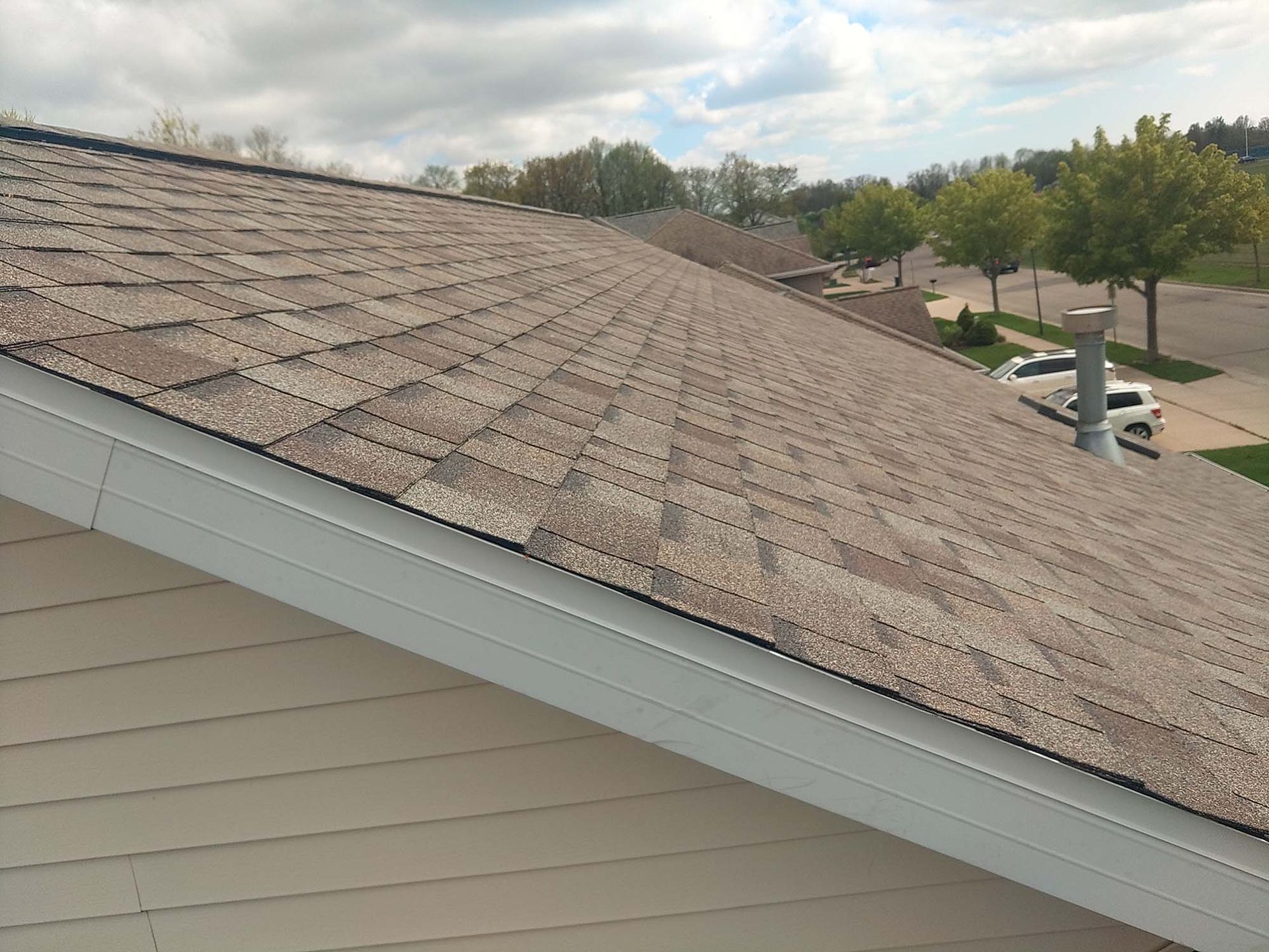 Roof with brown asphalt shingles on a cloudy day, white trim, and a glimpse of a street.