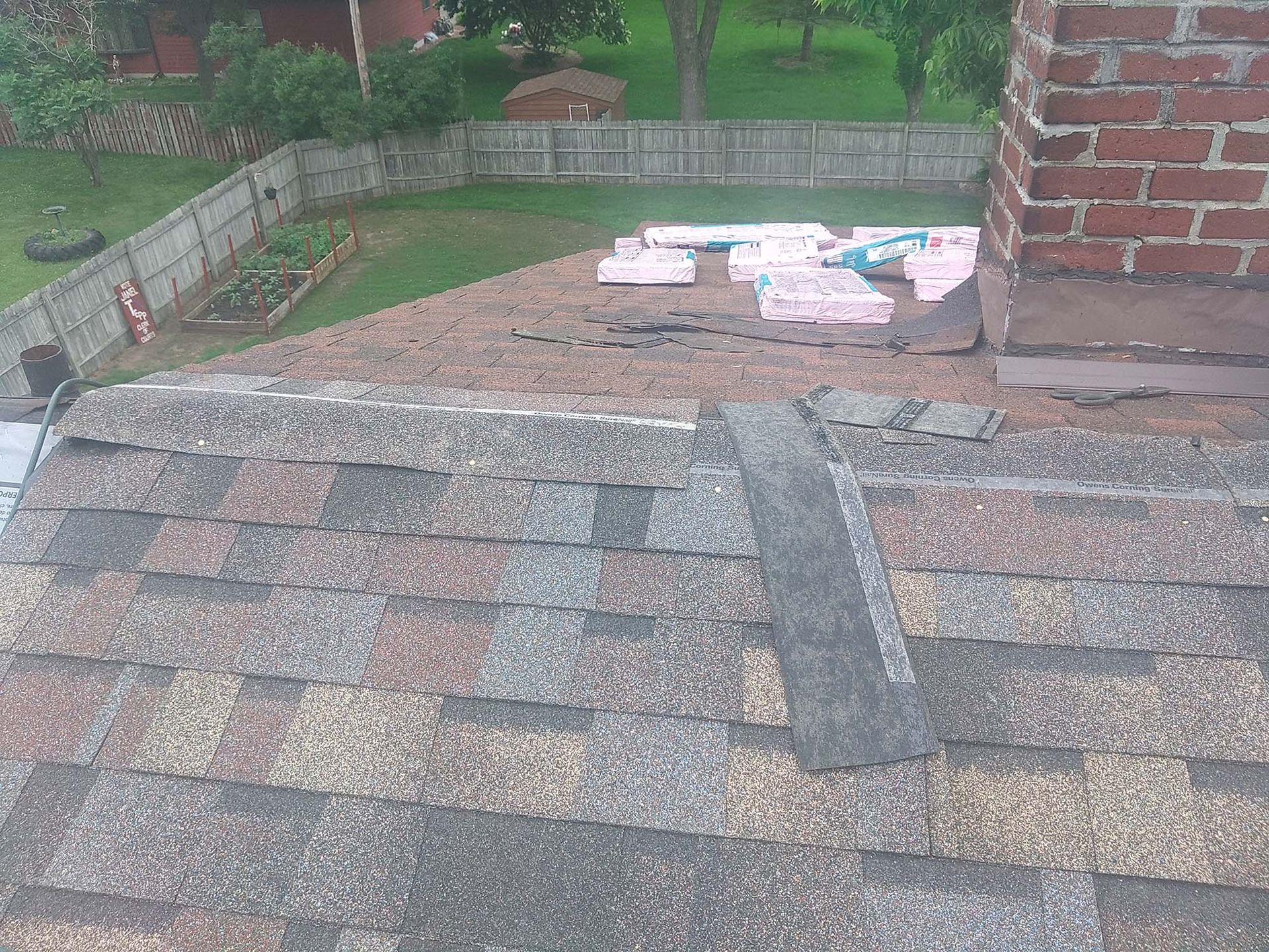 Damaged rooftop with exposed underlayment near a brick chimney, overlooking a backyard.