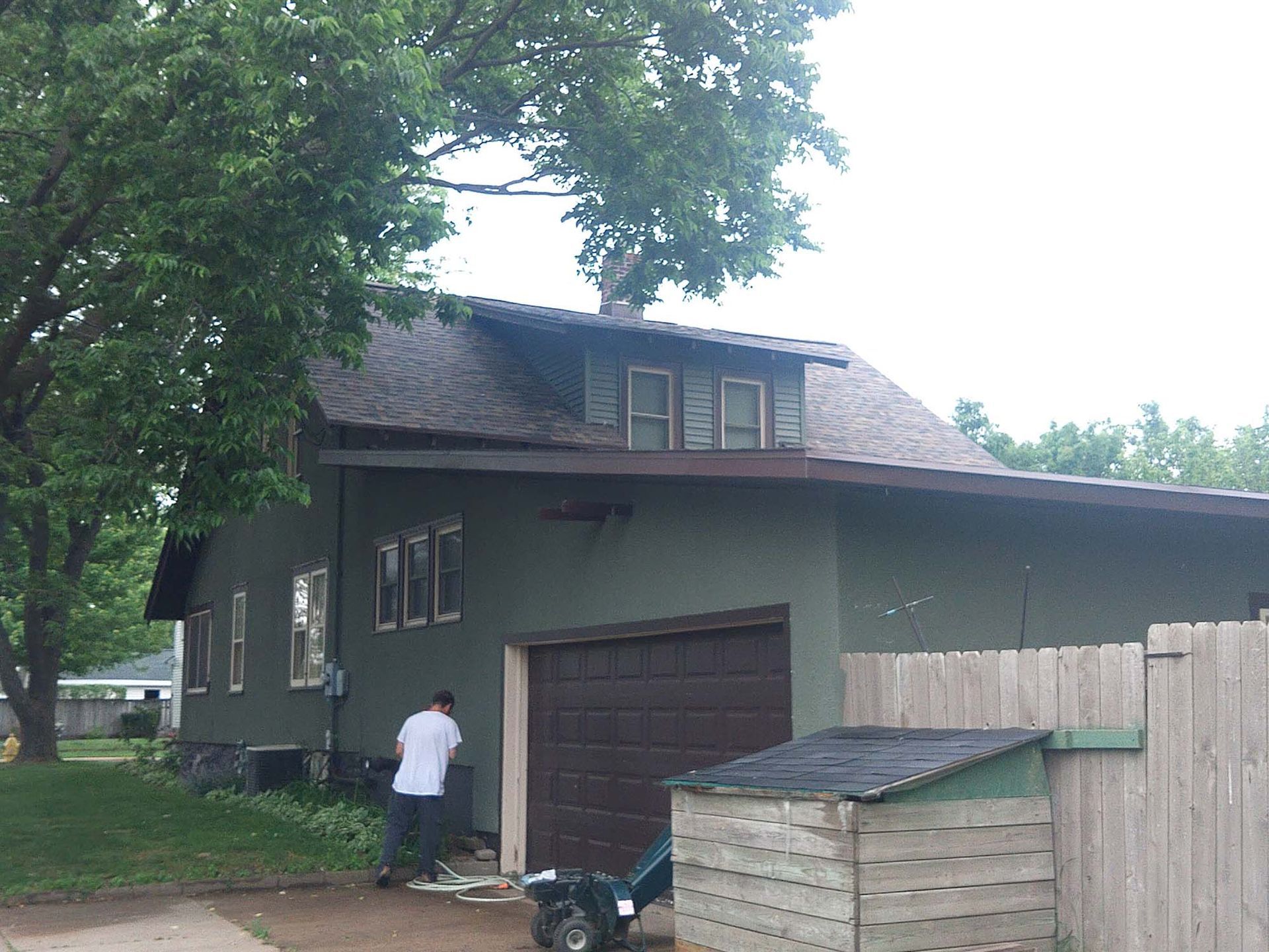 Green house with brown garage door, person working outside. Tree in background.