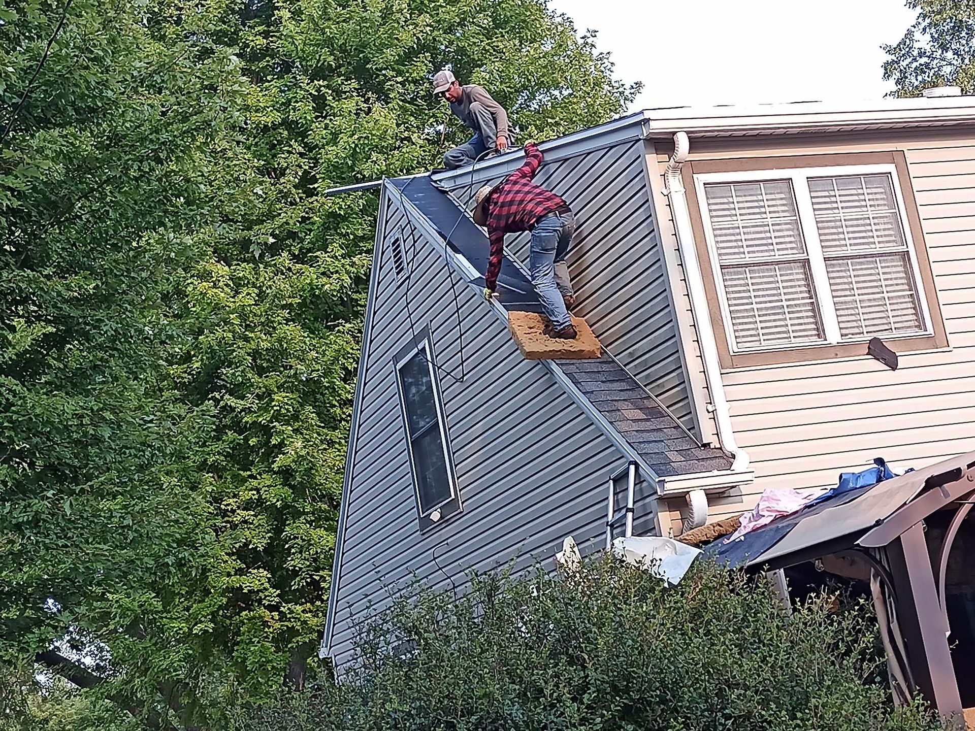 Three roofers working on a house's roof. One climbs, another steadies, and one stands at the peak.