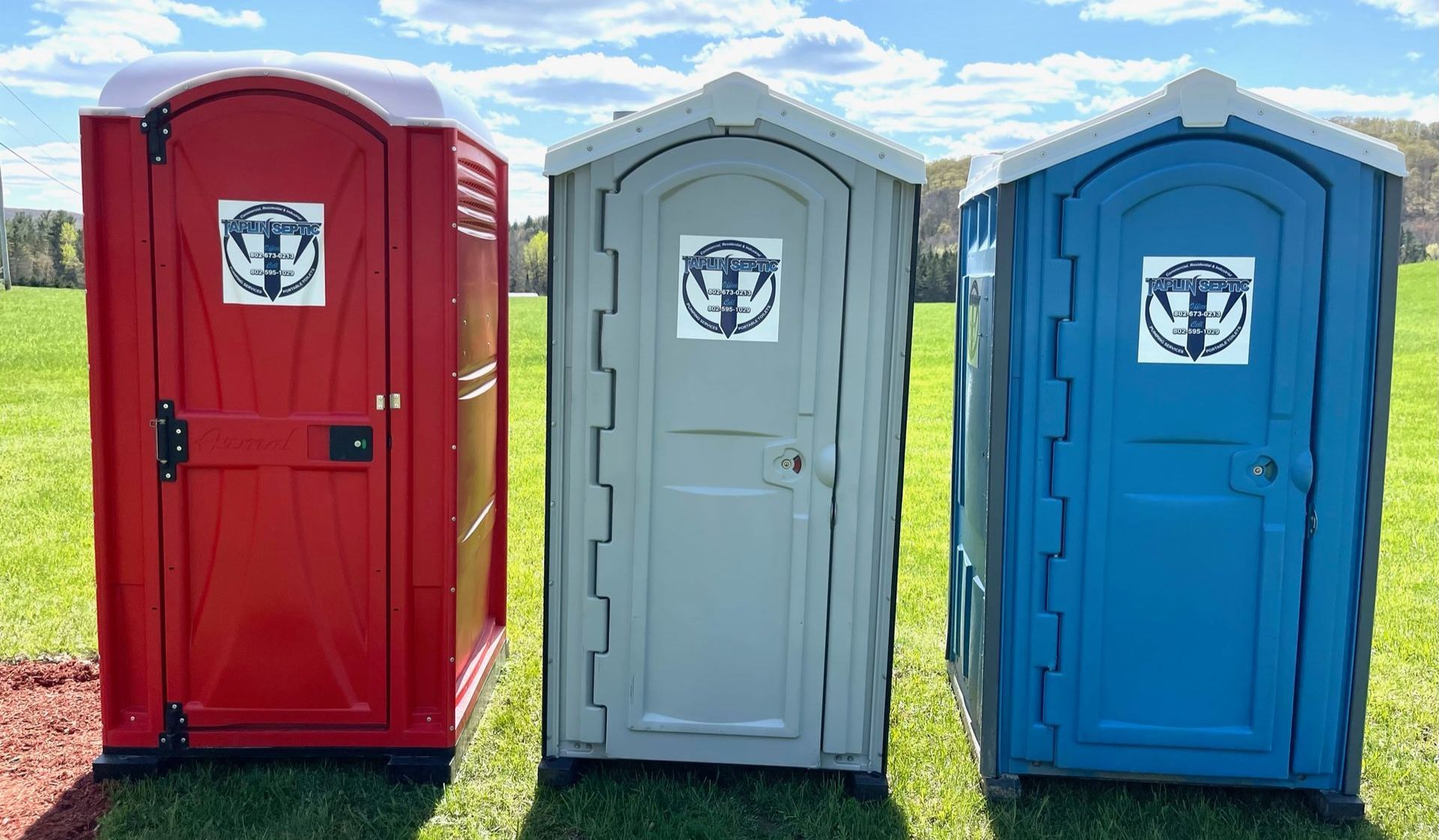 Red, white, and blue porta-potties in a line outside in the summer sun, showing a wide array of options available from Taplin Septic.