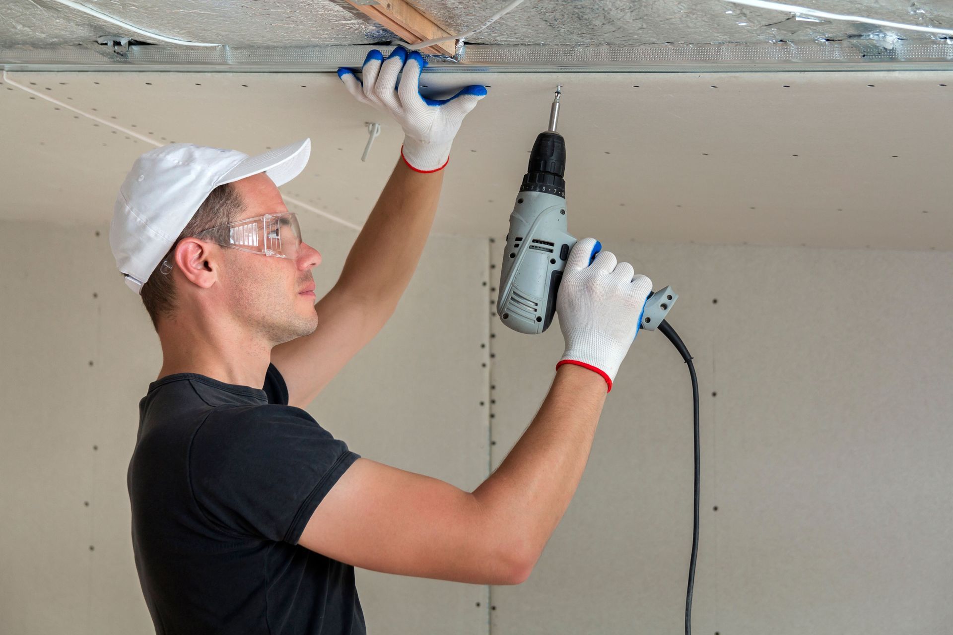 Man in white cap and safety glasses using a power drill to install drywall.