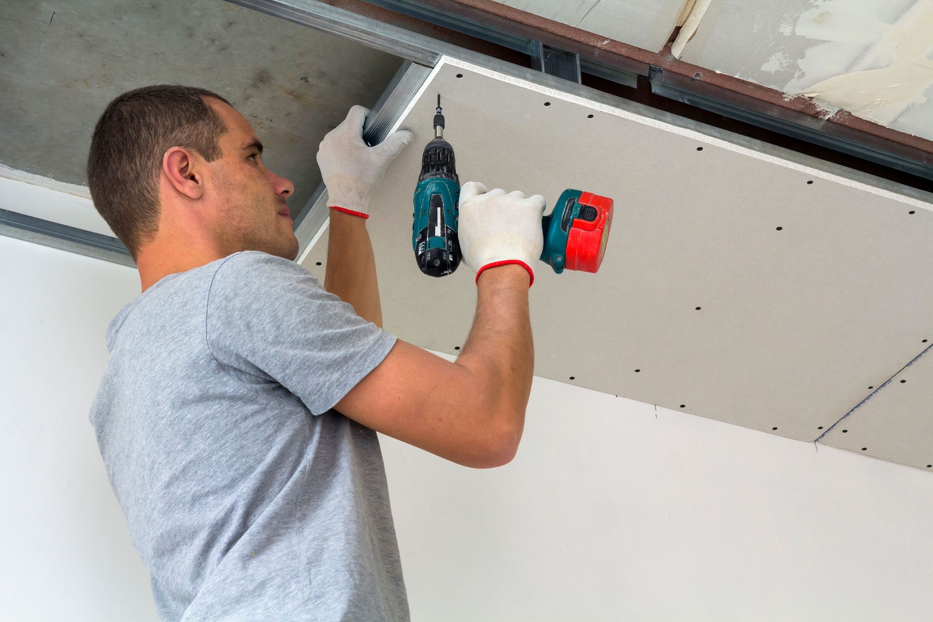 Man in a grey shirt and gloves using a drill to install drywall on a ceiling.
