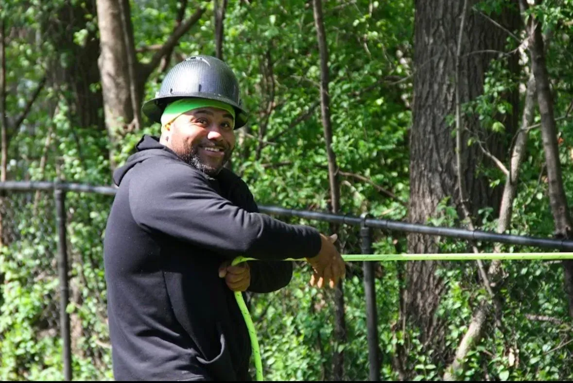Man in black hoodie and hard hat, smiling, holding a green rope in a wooded area.