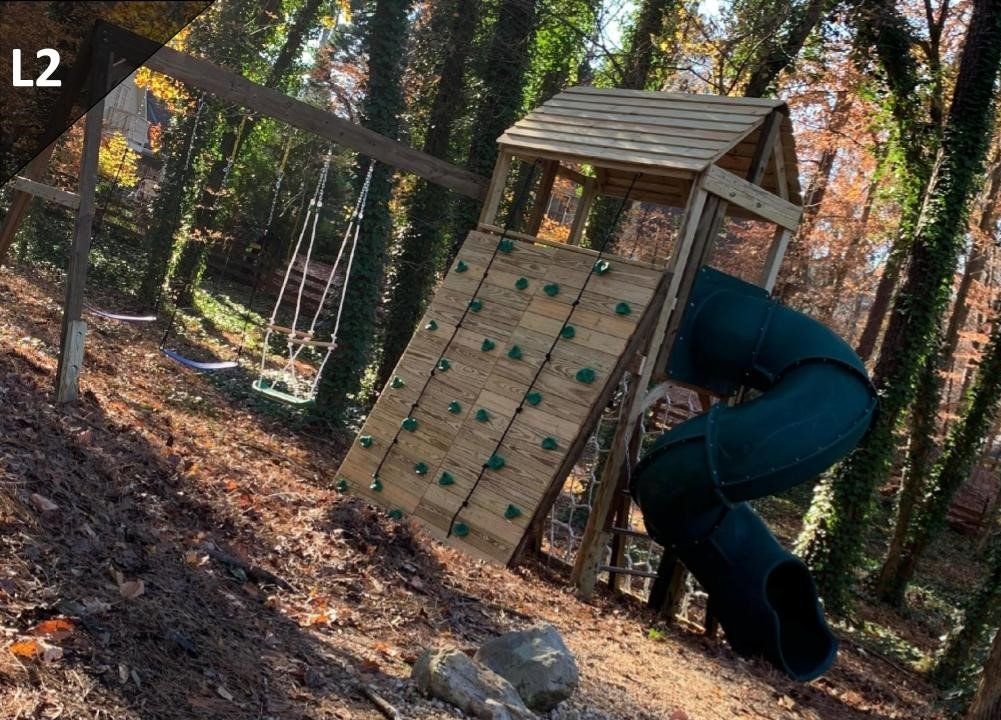 A wooden playground with a slide and a climbing wall in the woods.