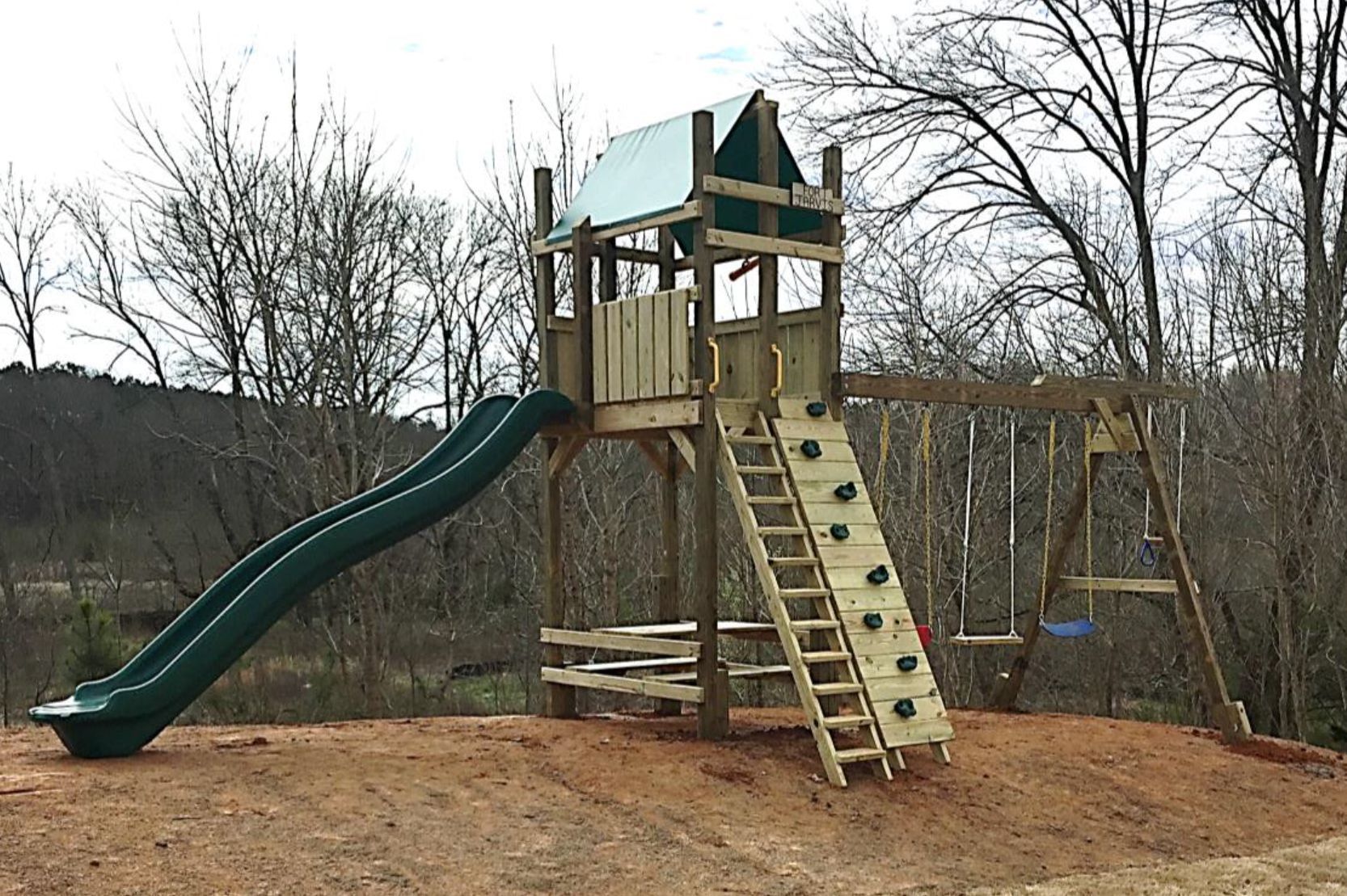 A wooden playground with a slide , swings , and climbing wall.