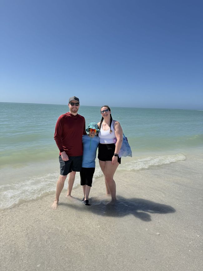 Bryant and his family are standing on a sandy beach with waves and a clear blue sky behind them.
