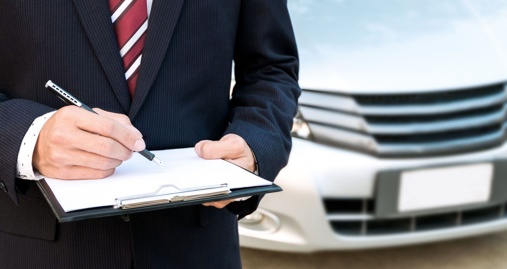 Businessperson writing on a clipboard in front of a car front grille