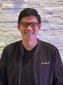 A man wearing glasses and a black jacket is smiling in front of a brick wall.