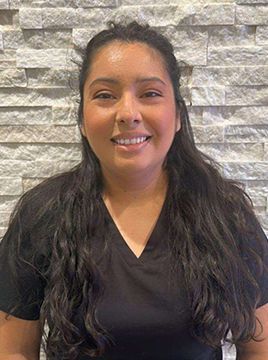 A woman in a black scrub top is smiling in front of a brick wall.