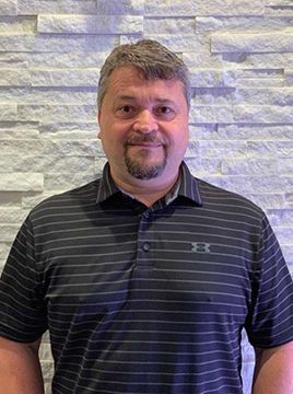 A man with a beard is wearing a striped shirt and standing in front of a brick wall.