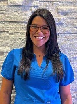 A woman wearing glasses and a blue scrub top is smiling in front of a brick wall.