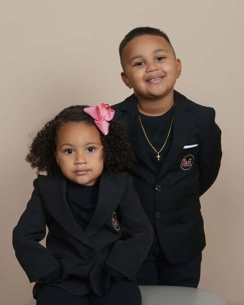 Two children in black outfits posing against a beige backdrop, one with a pink bow and both smiling.