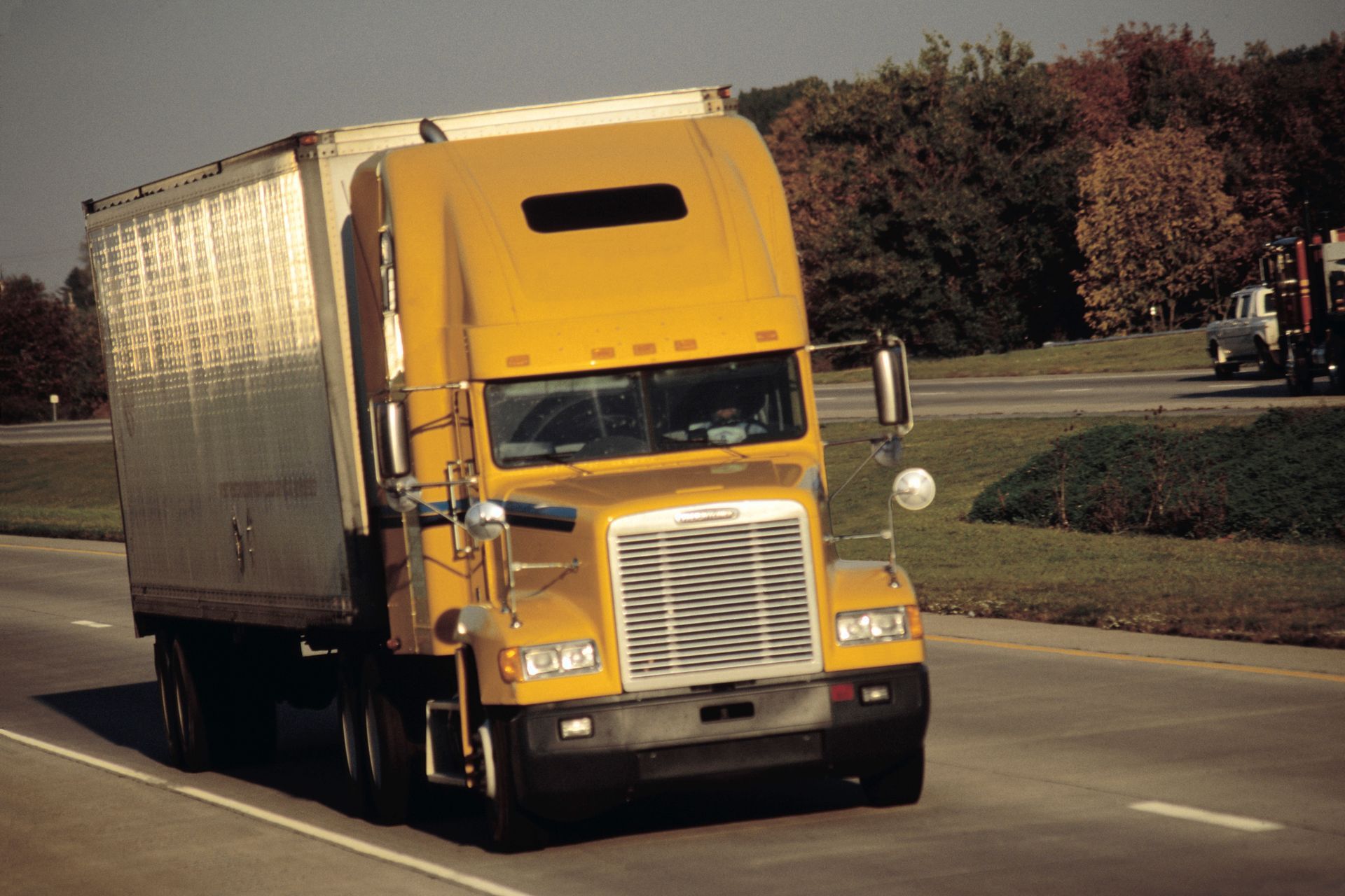 Yellow semi-truck driving on a road beside trees and a grassy median