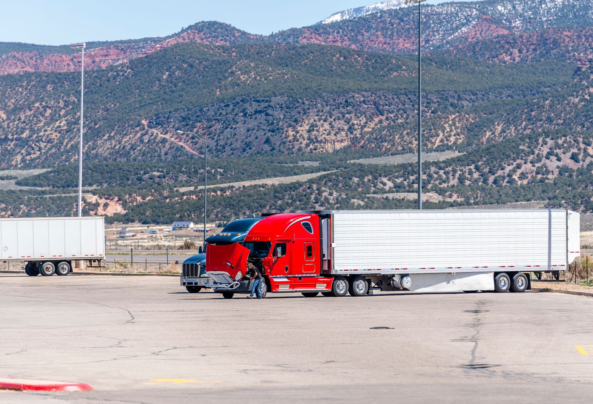 Red semi-truck with white trailer parked on a wide lot, mountains in the background