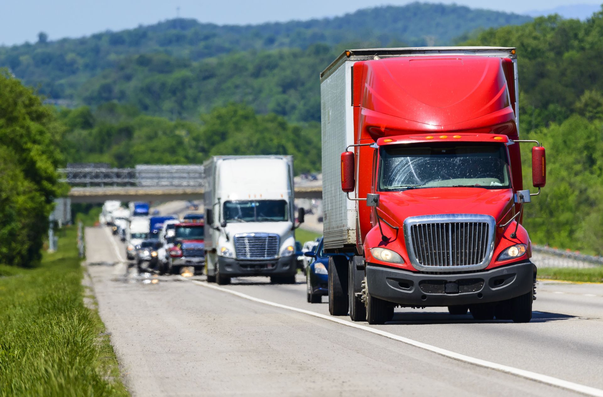 Red semi-truck leading a line of trucks on a highway beside green hills