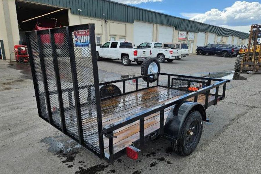 Black utility trailer with mesh ramp gate parked on wet pavement outside a warehouse
