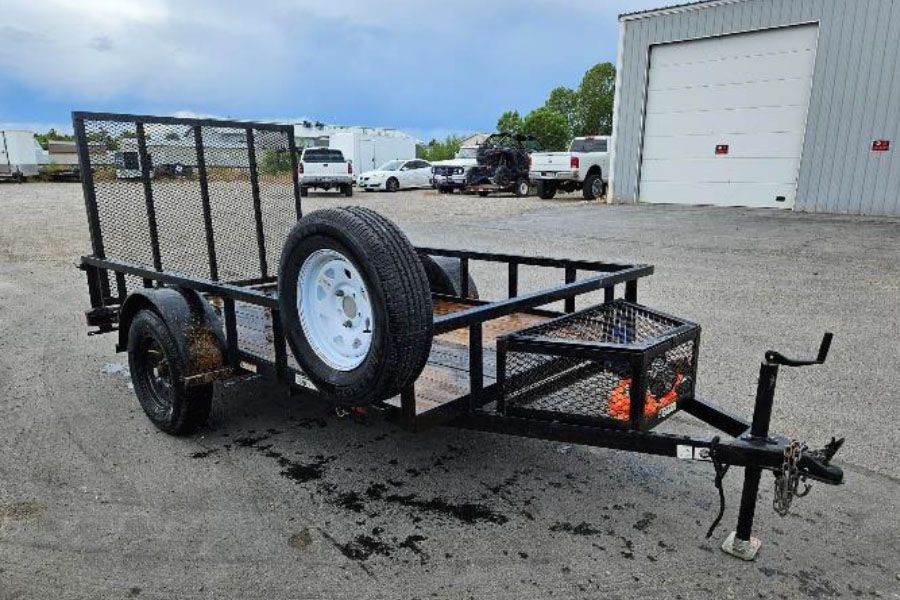 Black flatbed utility trailer with spare tire, parked on a gravel lot near warehouses.