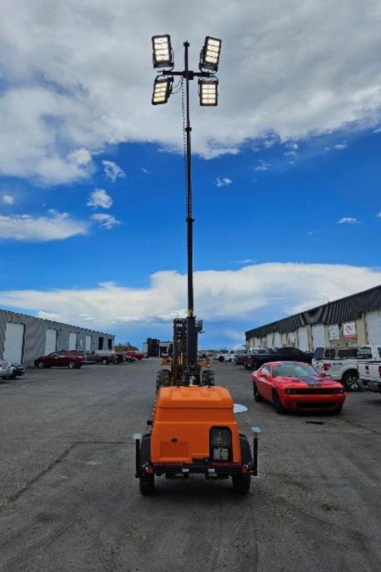 Orange mobile light tower on a lot between warehouses, with a red sports car nearby and blue sky overhead