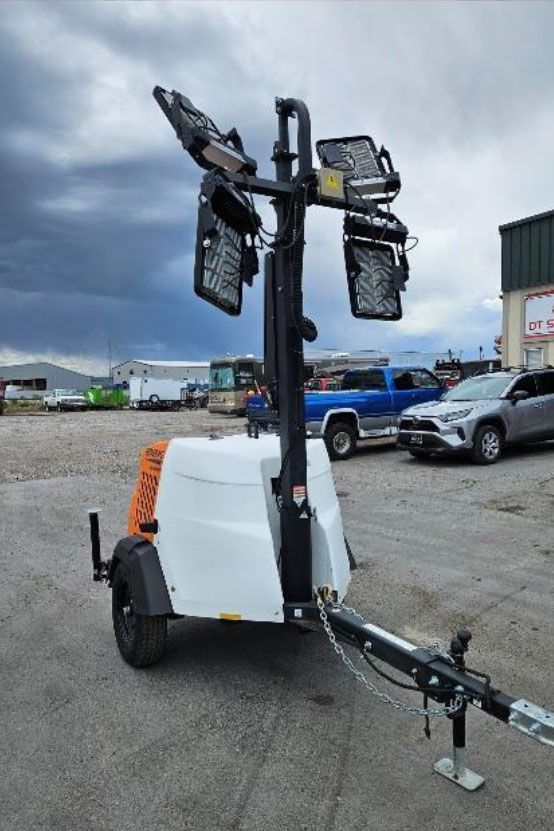 Portable light tower on a trailer in a gravel lot, with folded black lamps and a white base.
