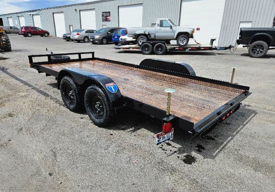 Rusty flatbed trailer with dual axles parked in an industrial lot beside warehouse buildings.