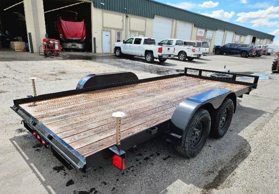 Flatbed utility trailer with weathered wooden deck and black rails, parked in an industrial lot with trucks nearby