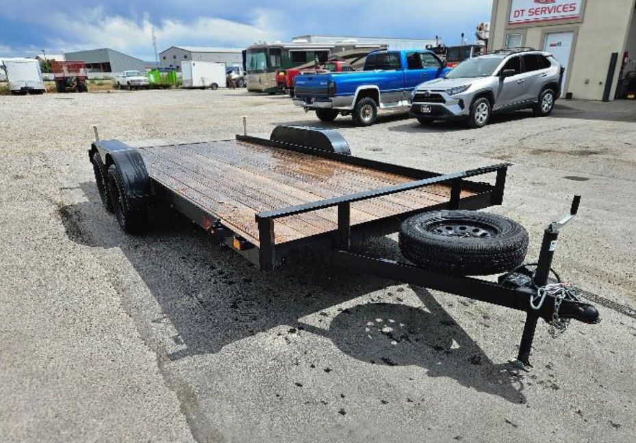 Flatbed trailer parked on gravel lot beside trucks and a service building