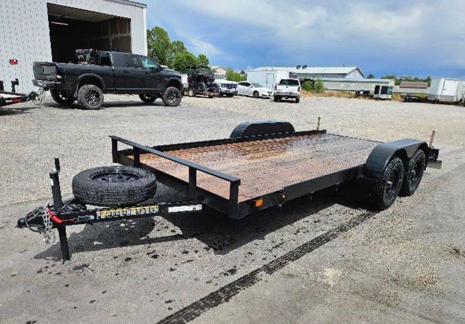 Black flatbed utility trailer parked on gravel with spare tire at front, industrial lot in background