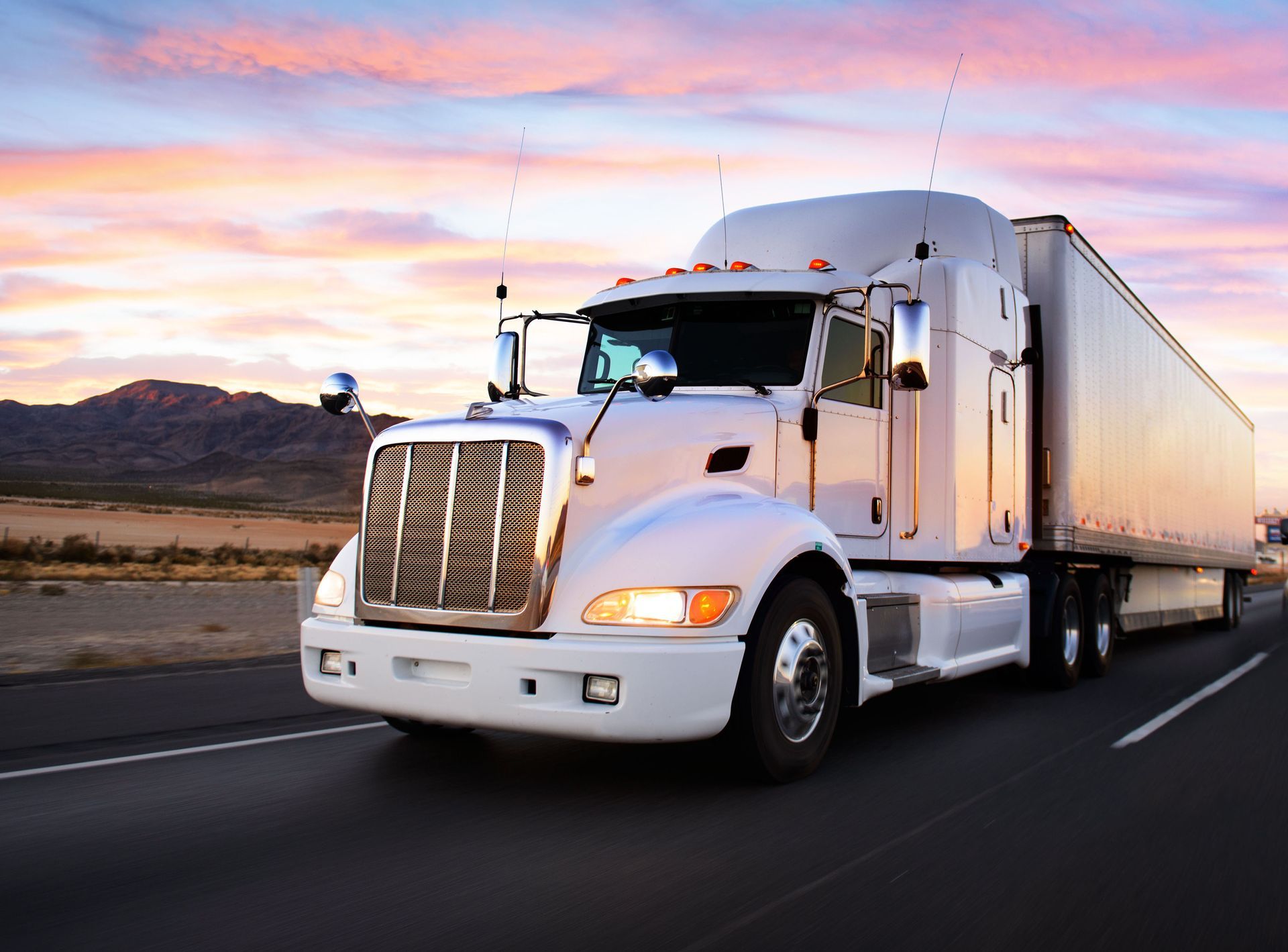 White semi-truck driving on a highway at sunset with mountains in the background