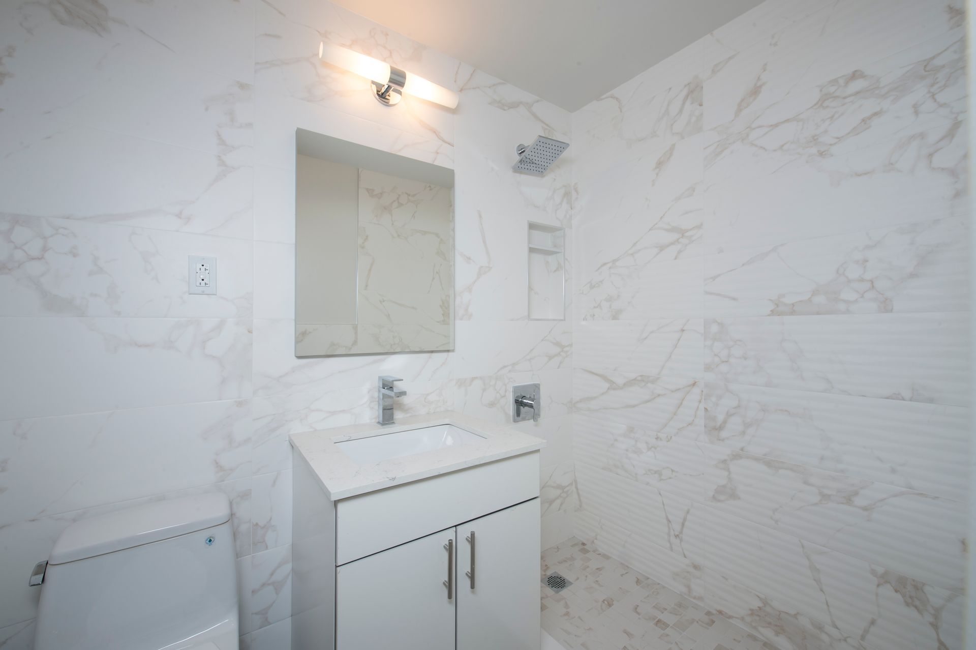 White bathroom with marble-patterned walls, vanity sink, mirror, and toilet light fixture on ceiling.