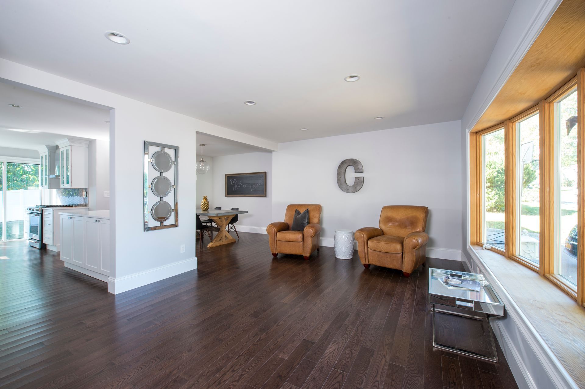 Bright living room with dark wood floors, tan chairs, wall art, and large windows overlooking greenery