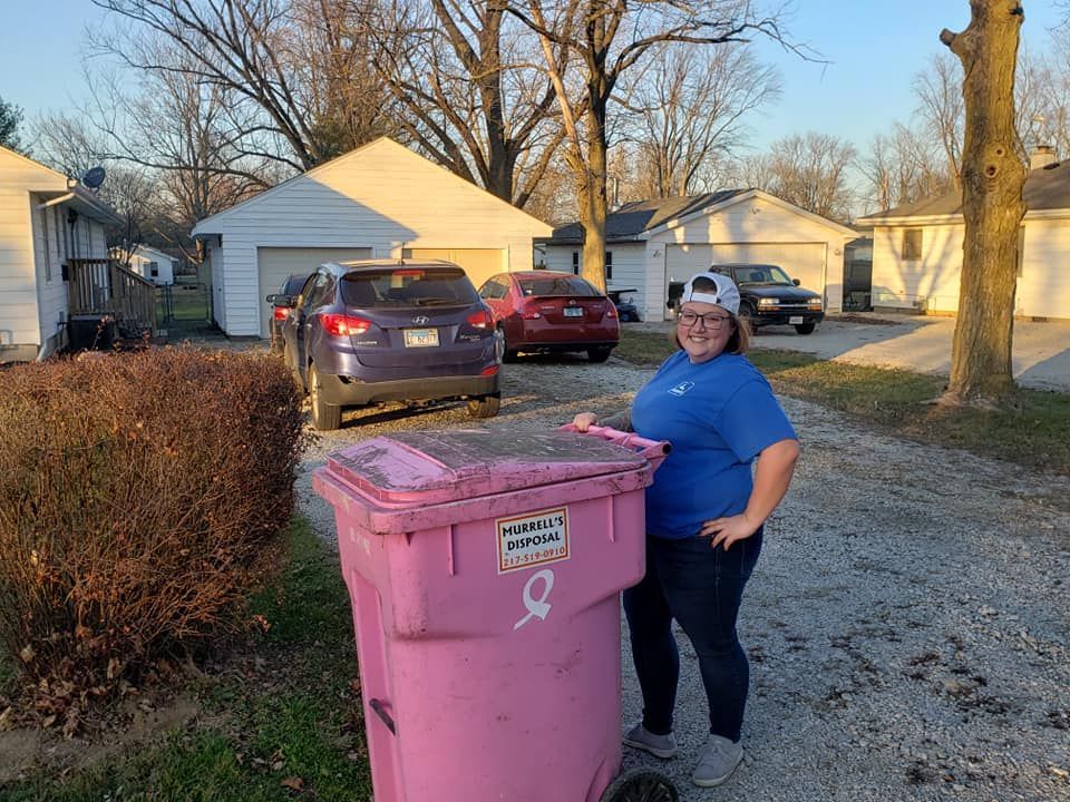 Woman in blue shirt and cap standing beside a pink trash bin, cars and houses in the background.