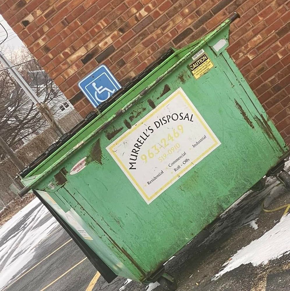 Green dumpster in a parking lot with a handicap sign and brick wall.