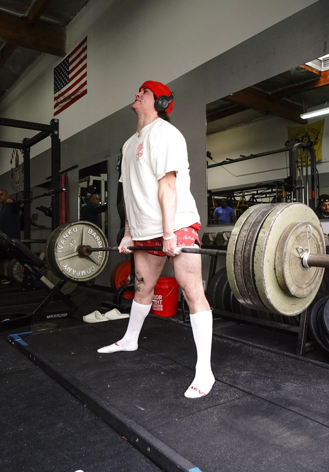Lifter performing a deadlift at a gym; barbell on the floor, red and white attire, American flag in background.