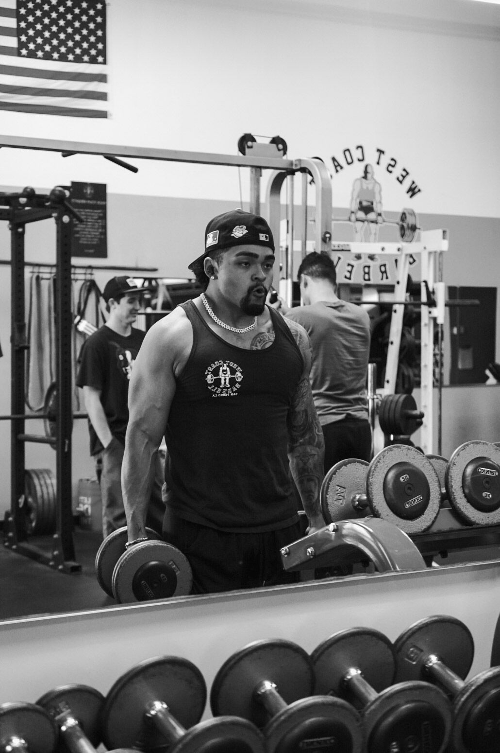 Man lifting dumbbells in a gym, looking in the mirror. Others are in the background, weight rack visible.