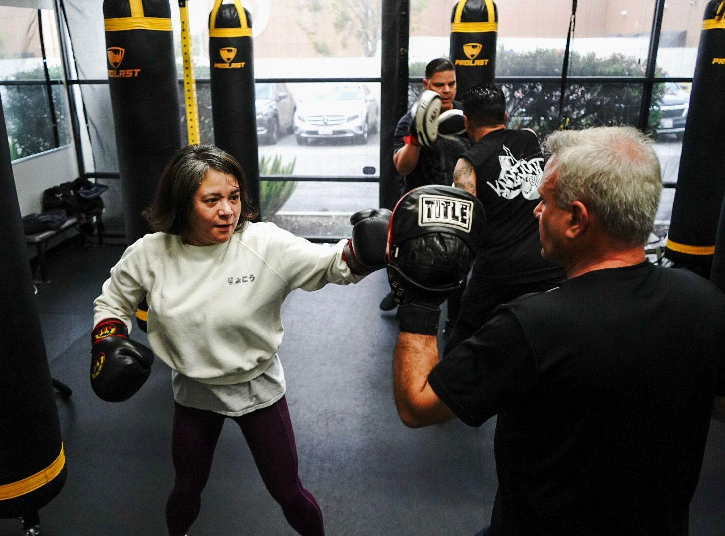 Woman throwing a punch in a boxing gym, training with a partner. Black punching bags are in the background.