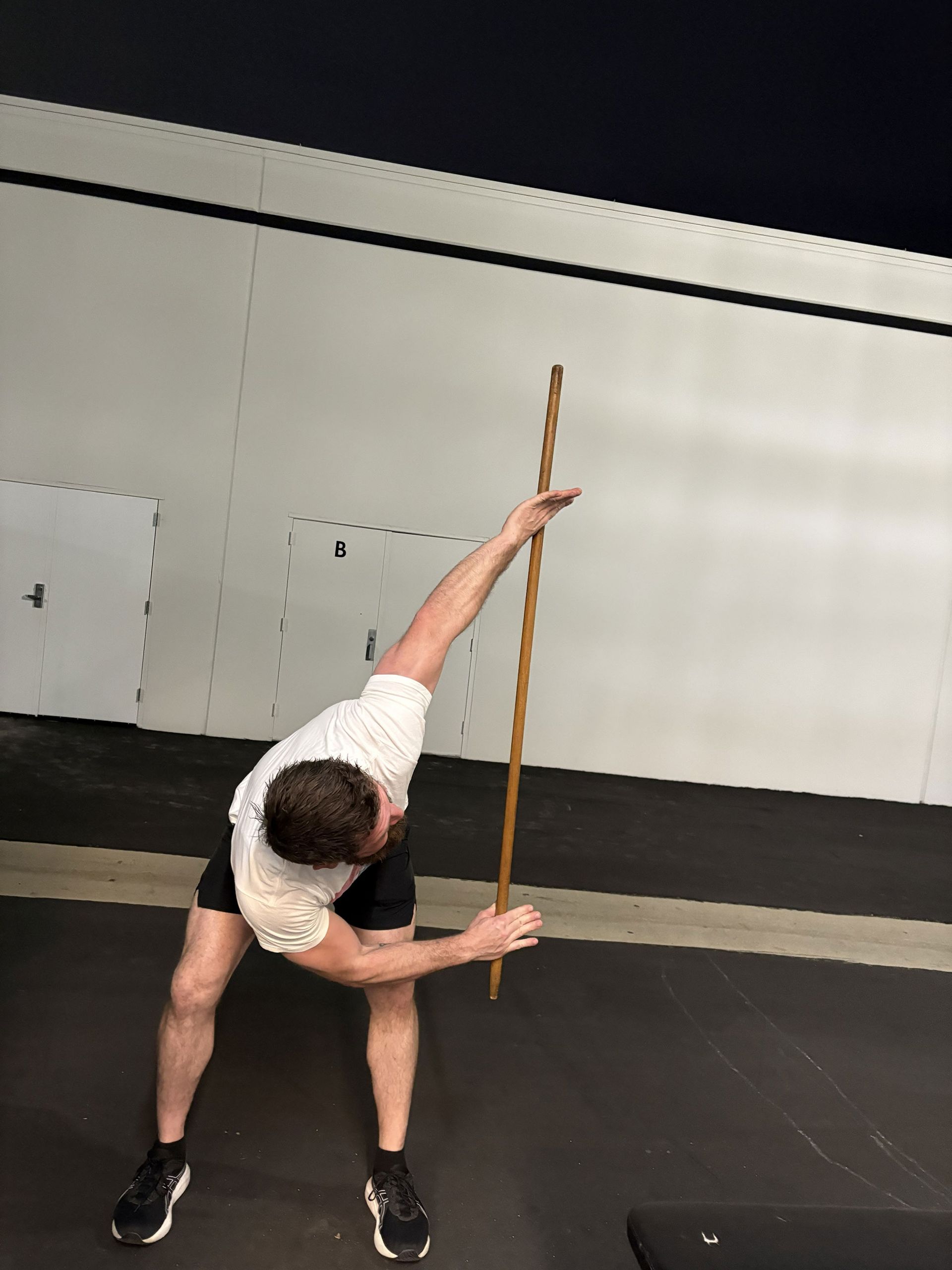 Man stretching with a long stick in front of a white wall. He is bent over and holding the stick.