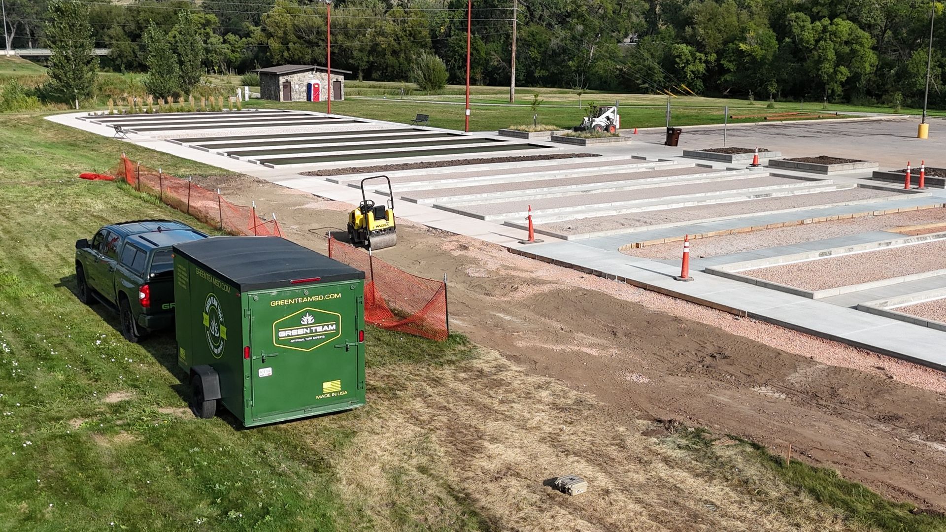Construction site: car pulling a green trailer, gravel and concrete pathways being built