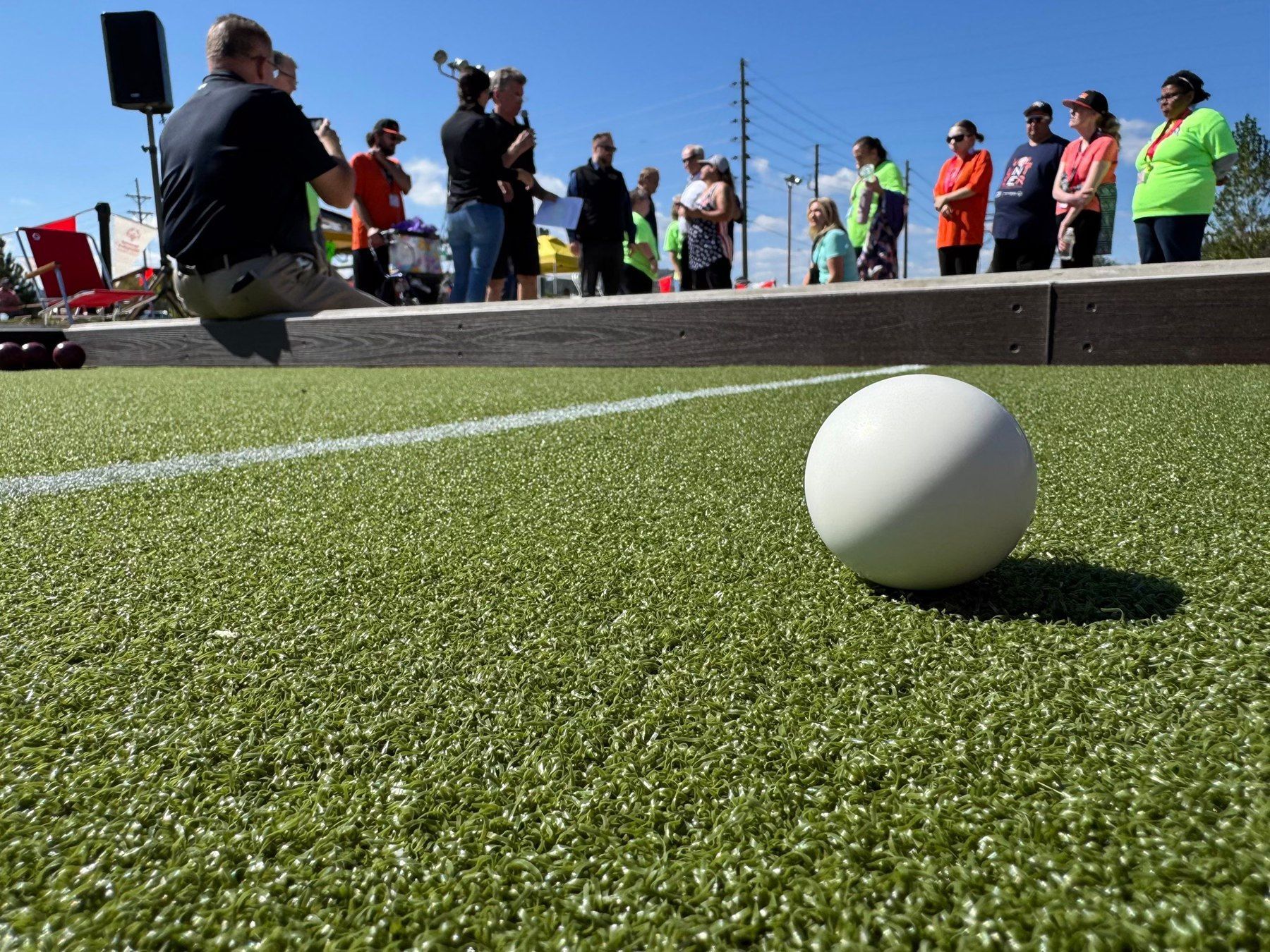 White bocce ball on green turf with people gathered on a raised platform, blue sky overhead